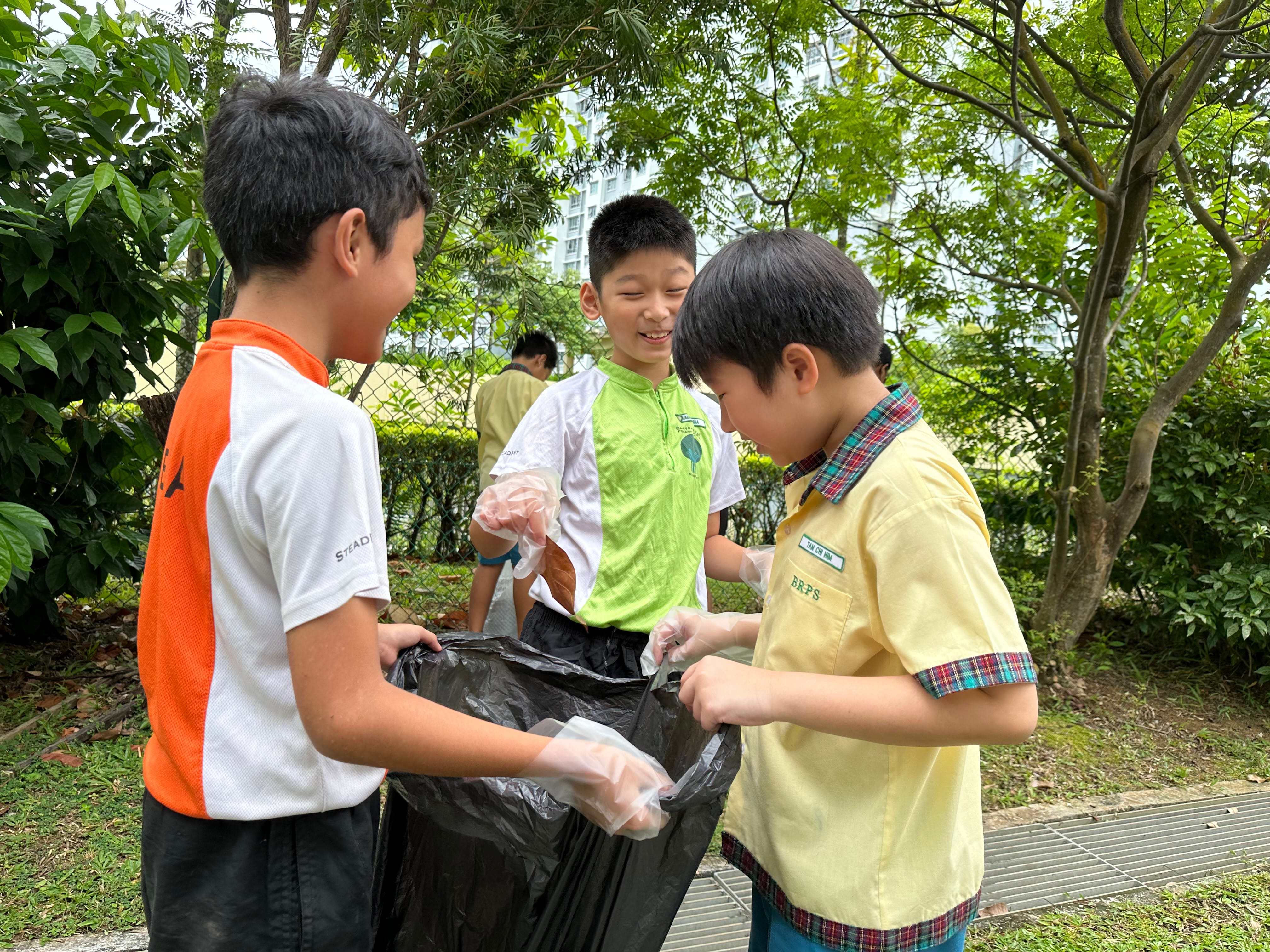 Three boys wearing gloves hold open a black trash bag outdoors under trees, smiling as they collect litter.